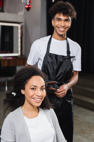 Positive african american client looking at camera near stylist with hair iron 