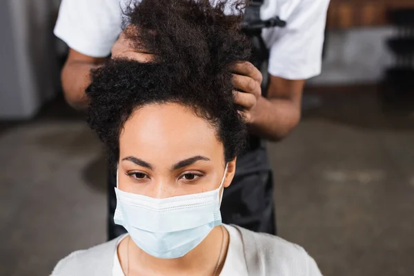若いですアフリカ系アメリカ人クライアントで医療マスク座っています近くヘアスタイリストオンぼやけた背景 — ストック写真