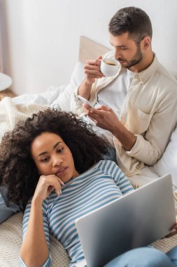 Afro-Amerikan bir kadının yatakta yattığını ve bilgisayarını akıllı telefonlu sevgilisinin yanında kahve içerken yüksek açılı görüntüsü.
