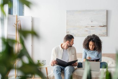 Elinde vergi dosyası olan sakallı bir adam ve yatağında oturan defterli Afrikalı Amerikalı kız arkadaşına bakıyor.