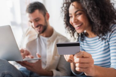credit card in hand on cheerful african american woman near boyfriend with laptop on blurred background clipart