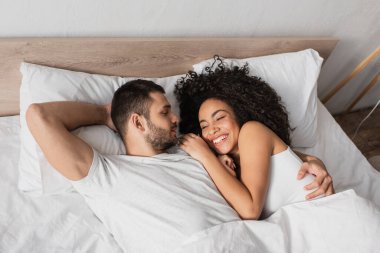 Yatakta gülümseyen Afrikalı Amerikalı kız arkadaşına bakan sakallı bir adamın yüksek açılı görüntüsü.