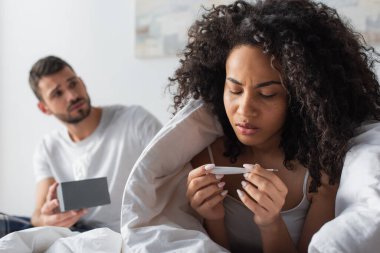 ill african american woman looking at digital thermometer near worried boyfriend on blurred background  clipart