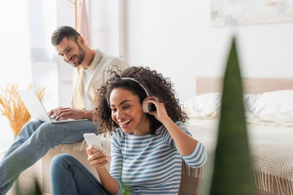幸せなアフリカ系アメリカ人女性ですワイヤレスヘッドフォンを保持スマートフォンの近くにボーイフレンドとノートパソコンを使用してぼやけた前景 — ストック写真
