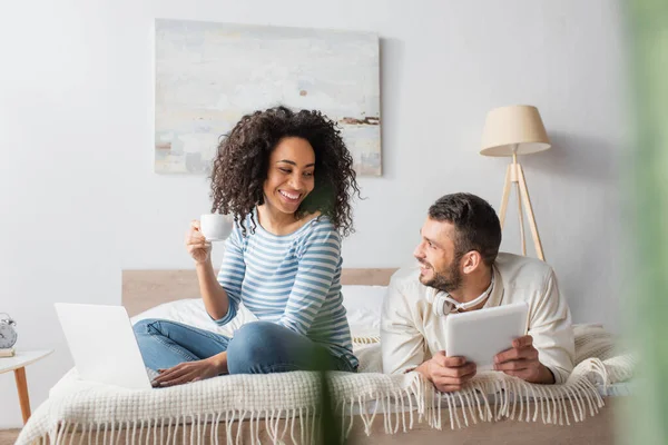 幸せなアフリカ系アメリカ人の女性がベッドに座ってカップを持ちデジタルタブレットでボーイフレンドの近くのノートパソコンを使って — ストック写真