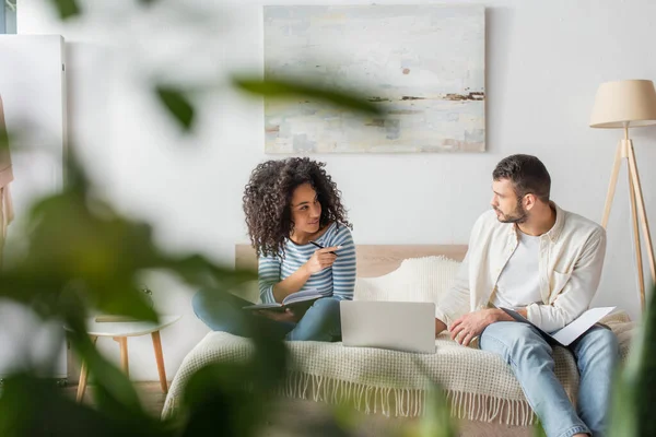 african american woman pointing at boyfriend sitting with folder near laptop on bed with blurred plant on foreground