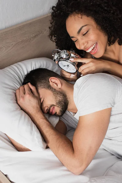 Cheerful African American Woman Holding Alarm Clock Displeased Man Sleeping — Stock Photo, Image