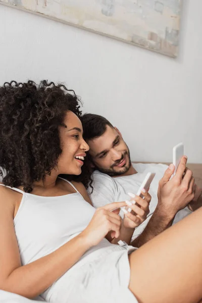 Cheerful Interracial Couple Using Cellphones Bedroom — Stockfoto