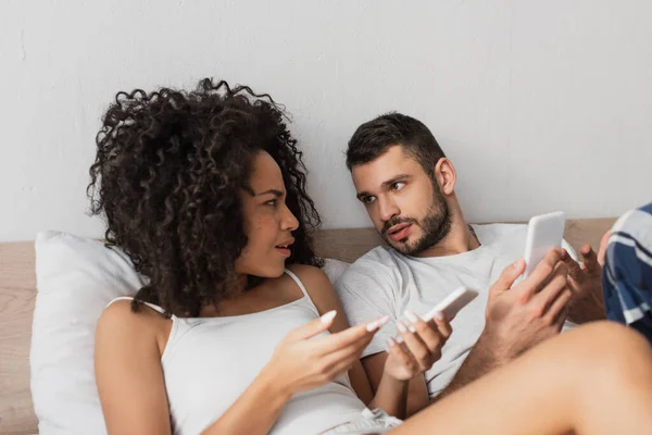 Pareja Interracial Disgustado Sosteniendo Los Teléfonos Celulares Mirándose Dormitorio —  Fotos de Stock