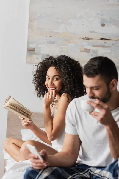 Cheerful African American Woman Holding Book Looking Boyfriend Holding Smartphone — Stock Photo, Image