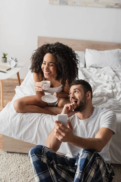 幸せなアフリカ系アメリカ人女性保持カップとともにコーヒー髭のボーイフレンド使用してスマートフォンでベッドルーム — ストック写真