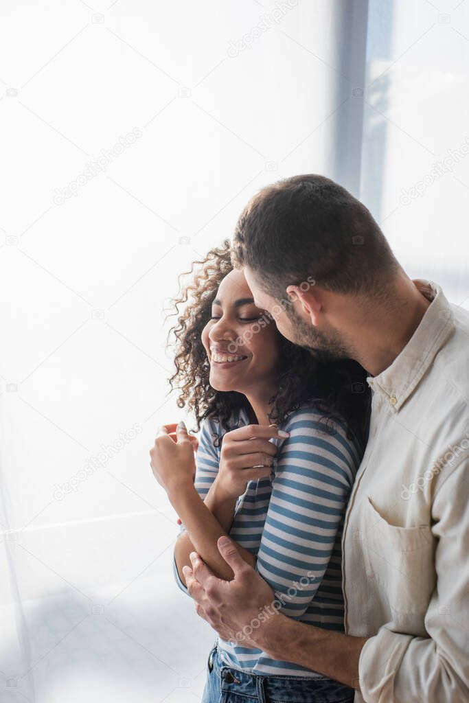 cheerful multiethnic couple smiling while hugging at home