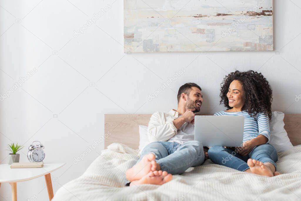 happy interracial couple chilling on bed and watching movie on laptop 
