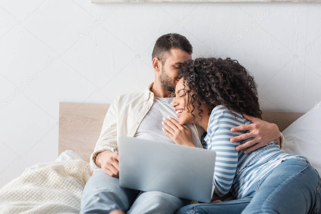 man kissing african american girlfriend while chilling on bed with laptop 