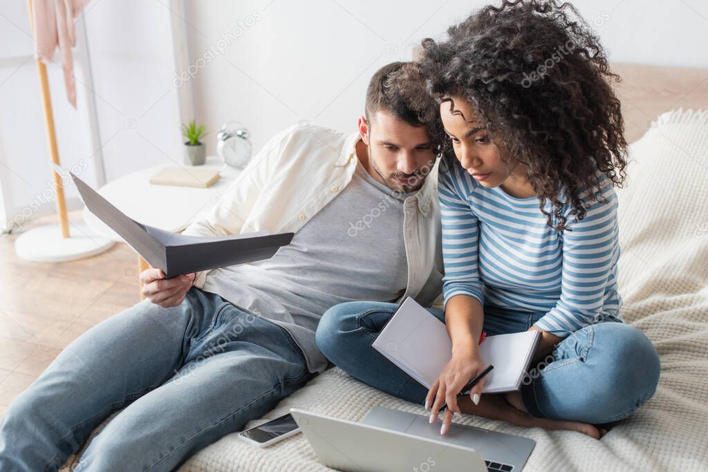 african american woman with notebook using laptop near bearded boyfriend holding folder in bedroom