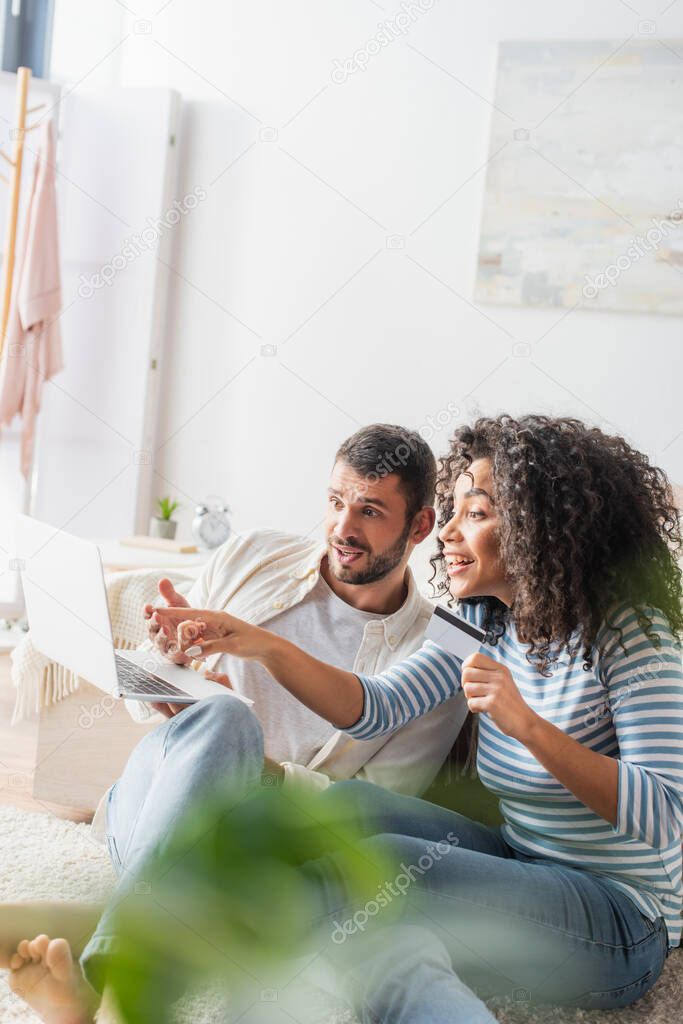 excited interracial couple shopping online while using laptop in bedroom with blurred foreground 