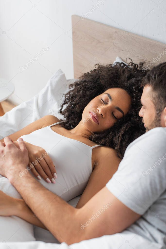 blurred bearded man looking at african american girlfriend sleeping on bed