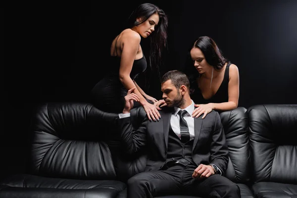 passionate women seducing man in suit sitting on leather couch isolated on black