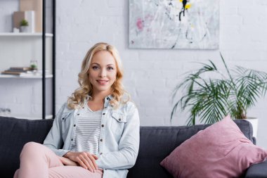 Kanepede yastığın yanındaki kameraya bakan gülümseyen kadın. 