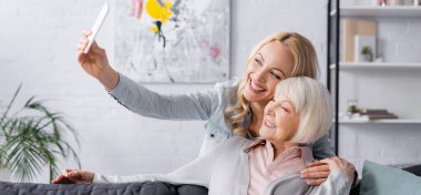 Gülümseyen yaşlı kadın kızının yanında selfie çekiyor, afiş. 