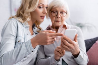 Geçmişi bulanık bir kadın parmaklarıyla yaşlı annenin elindeki akıllı telefonu işaret ediyor. 
