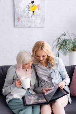 Smiling woman looking at photo album near elderly mother with cup on couch  clipart
