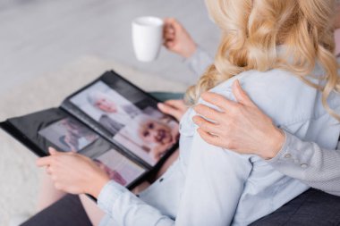 Cropped view of senior mother with cup hugging daughter with blurred photo album  clipart