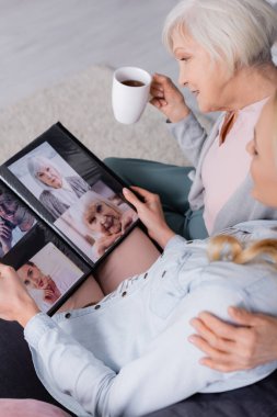 Elinde fincanla gülümseyen yaşlı kadın albümde bulanık kızının fotoğraflarına bakıyor. 