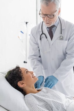 Plastik eldivenli olgun bir doktor. Klinikteki gülümseyen Afrikalı Amerikalı kadının omzunu inceliyor.