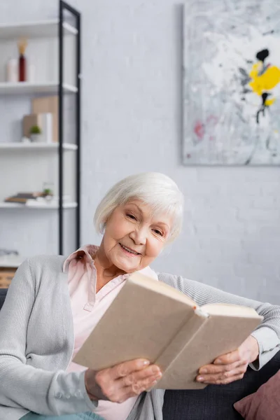 Glimlachende Oudere Vrouw Leest Boek Wazig Voorgrond Thuis — Stockfoto