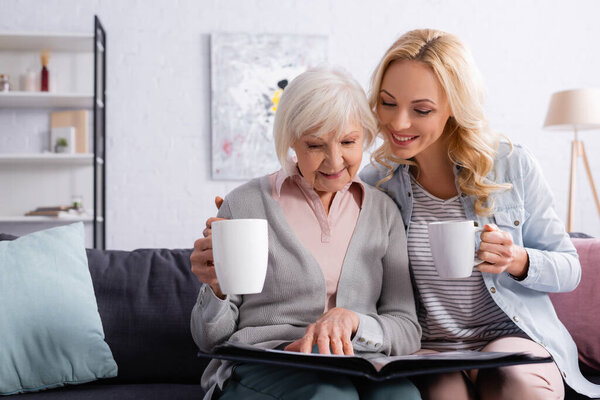 Улыбающаяся пожилая женщина с чашкой, глядя на фотоальбом рядом с дочерью 