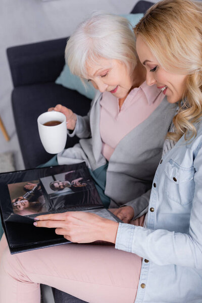 Smiling woman looking at photos in album near senior parent with tea 