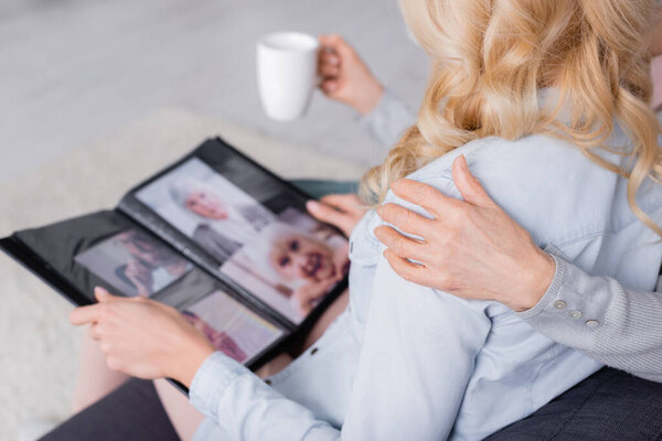 Cropped view of senior mother with cup hugging daughter with blurred photo album 