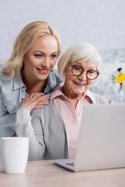 Mujer Alegre Abrazando Madre Mayor Cerca Computadora Portátil Taza Primer — Foto de Stock