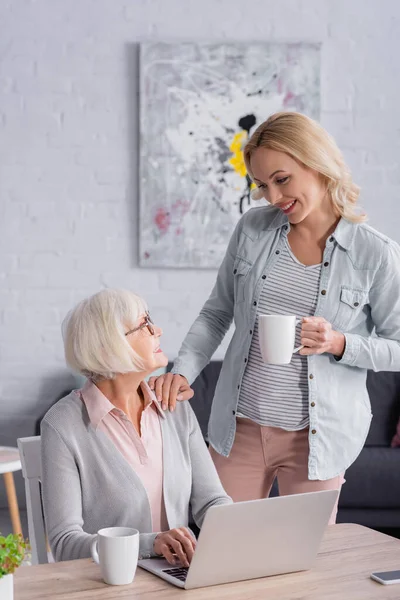 Mujer Sonriente Sosteniendo Taza Cerca Madre Mayor Utilizando Ordenador Portátil — Foto de Stock