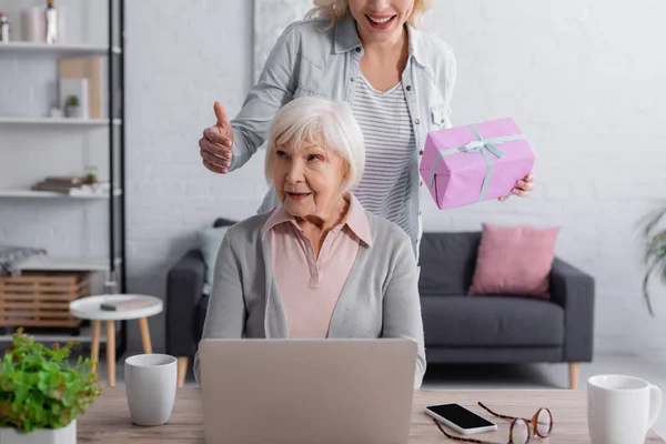 Mujer Sonriente Sosteniendo Presente Cerca Madre Anciana Utilizando Ordenador Portátil — Foto de Stock