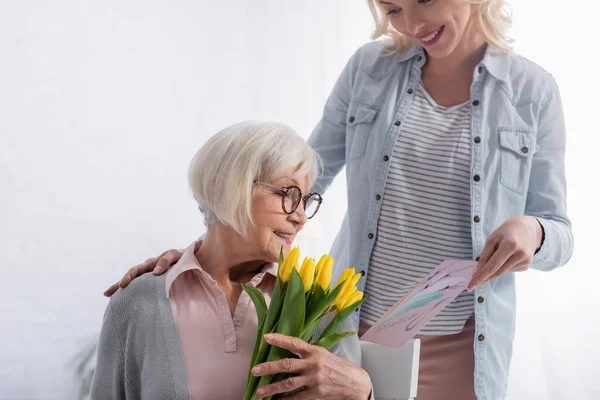 Mujer Sonriente Sosteniendo Tarjeta Felicitación Cerca Madre Anciana Con Tulipanes —  Fotos de Stock