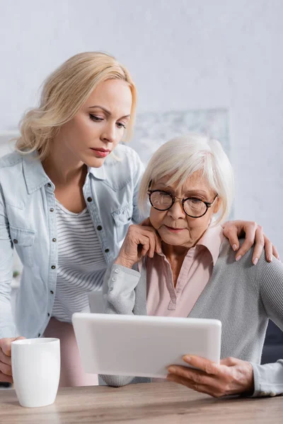 Mujer Abrazando Anciana Madre Con Tableta Digital Cerca Taza Mesa — Foto de Stock