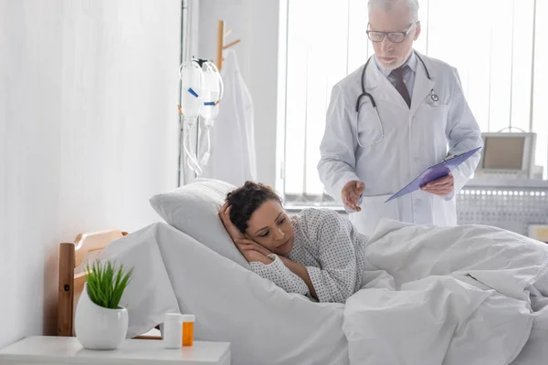 中年医師クリップボードスタンド近く動揺アフリカ系アメリカ人女性が病院のベッドに横たわっています — ストック写真