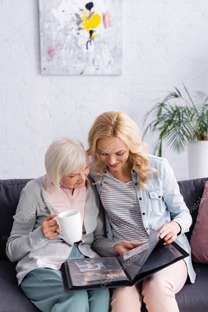 Smiling woman looking at photo album near elderly mother with cup on couch 