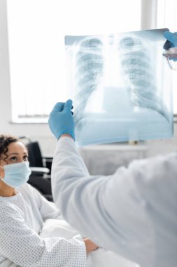lungs x-ray in hands of doctor in latex gloves near african american woman in medical mask on blurred background clipart
