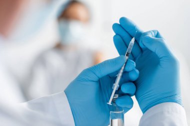 Ellerinde şırınga ve şişe tutan lateks eldivenli bir doktor. Arkaplanı bulanık olan Afrikalı Amerikalı bir kadının yanında aşı taşıyor.