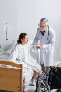 Doktor, Afro-Amerikan kadının hastanede tekerlekli sandalyenin yanından kalkmasına yardım ediyor.