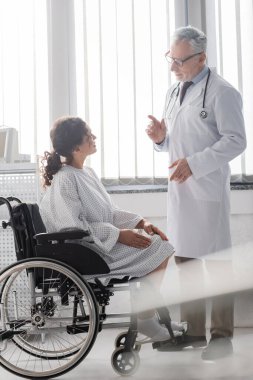 Olgun doktor, siyah önplanda tekerlekli sandalyede oturan Afrikalı Amerikalı kadınla konuşuyor.