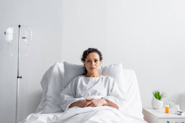 Hastalıklı Afro-Amerikalı kadın hastane yatağında otururken kameraya bakıyor.