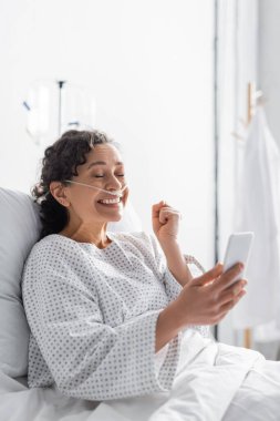 excited african american woman showing win gesture while holding cellphone in clinic clipart