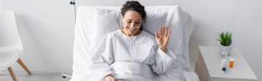 cheerful african american woman waving hand while sitting in hospital bed, banner clipart