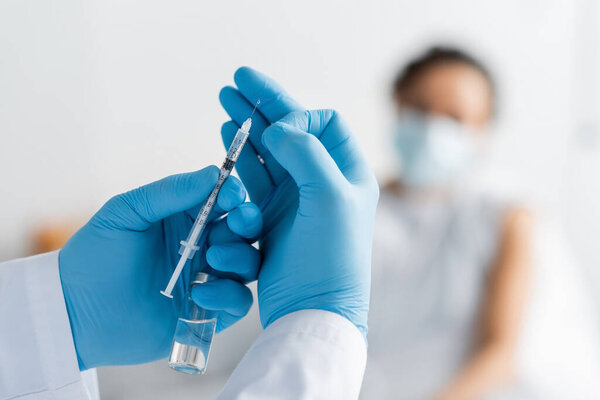 blurred african american patient near doctor in latex gloves holding bottle and syringe with vaccine
