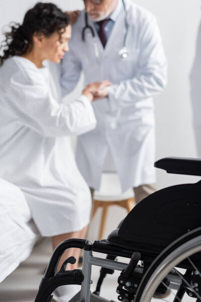 selective focus of wheelchair near doctor helping african american woman getting up from bed in clinic