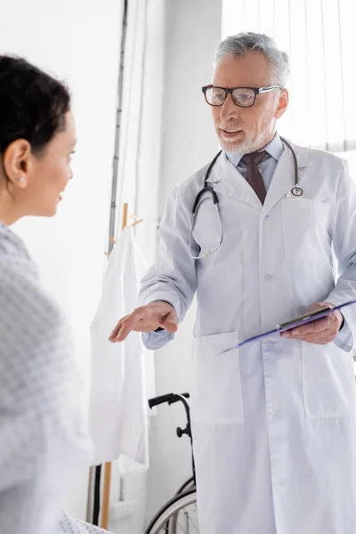 mature doctor with clipboard pointing with hand while talking to african american woman on blurred foreground